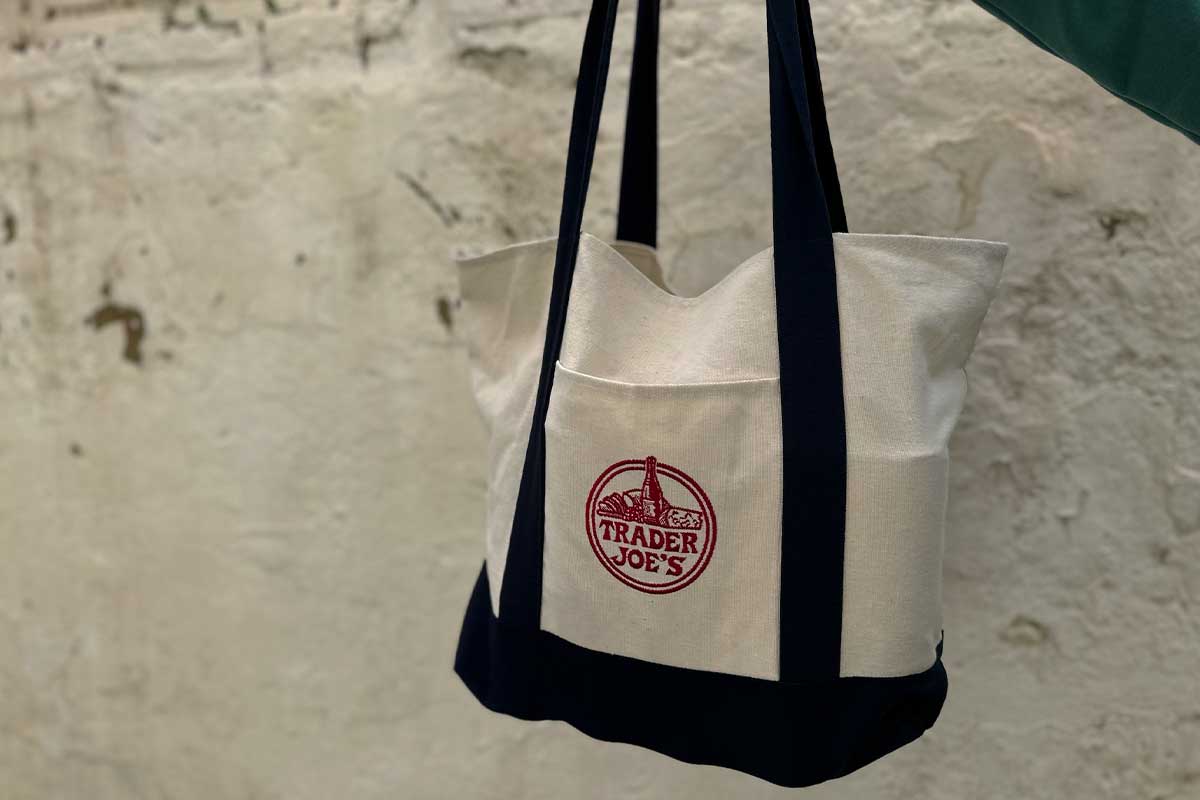 Borsa shopper di tela Trader Joe's bianca e blu con logo rosso appesa a una parete chiara